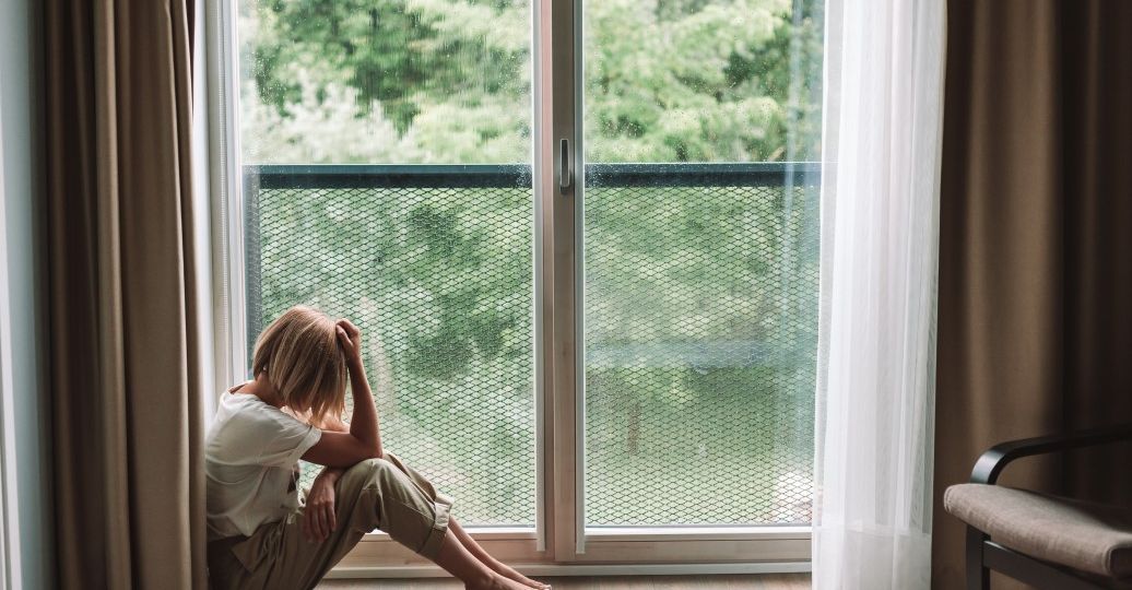 sad-woman-sitting-on-a-floor-looking-through-the-window-its-raining-outside_t20_dzpg0o