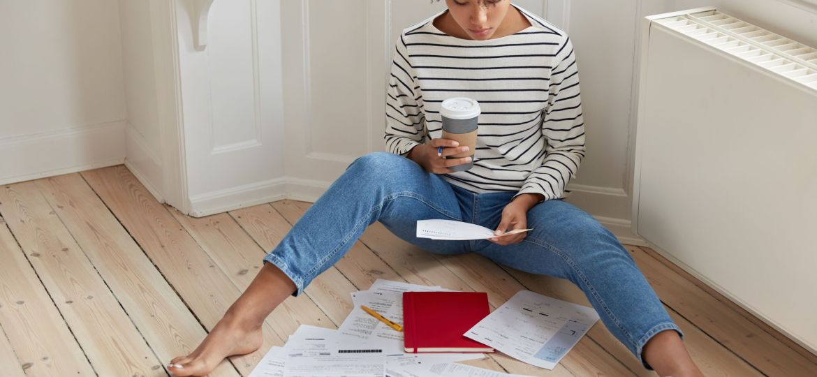 People, work and ethnicity concept. Dark skinned manager tries to complete marketing task, reads attentively documentation, holds paper and takeaway coffee, uses modern gadget for searching online