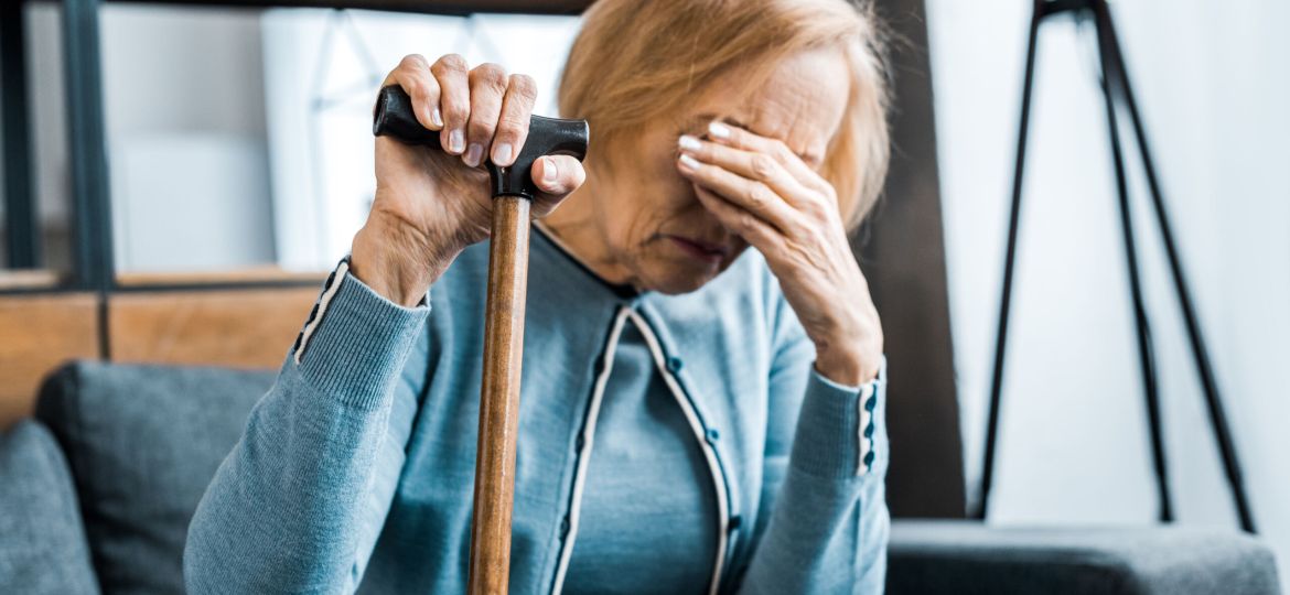 upset senior woman holding walking stick, covering face with hand and crying at home